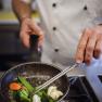 A cook in a white uniform fries vegetables in a pan on a gas stove.