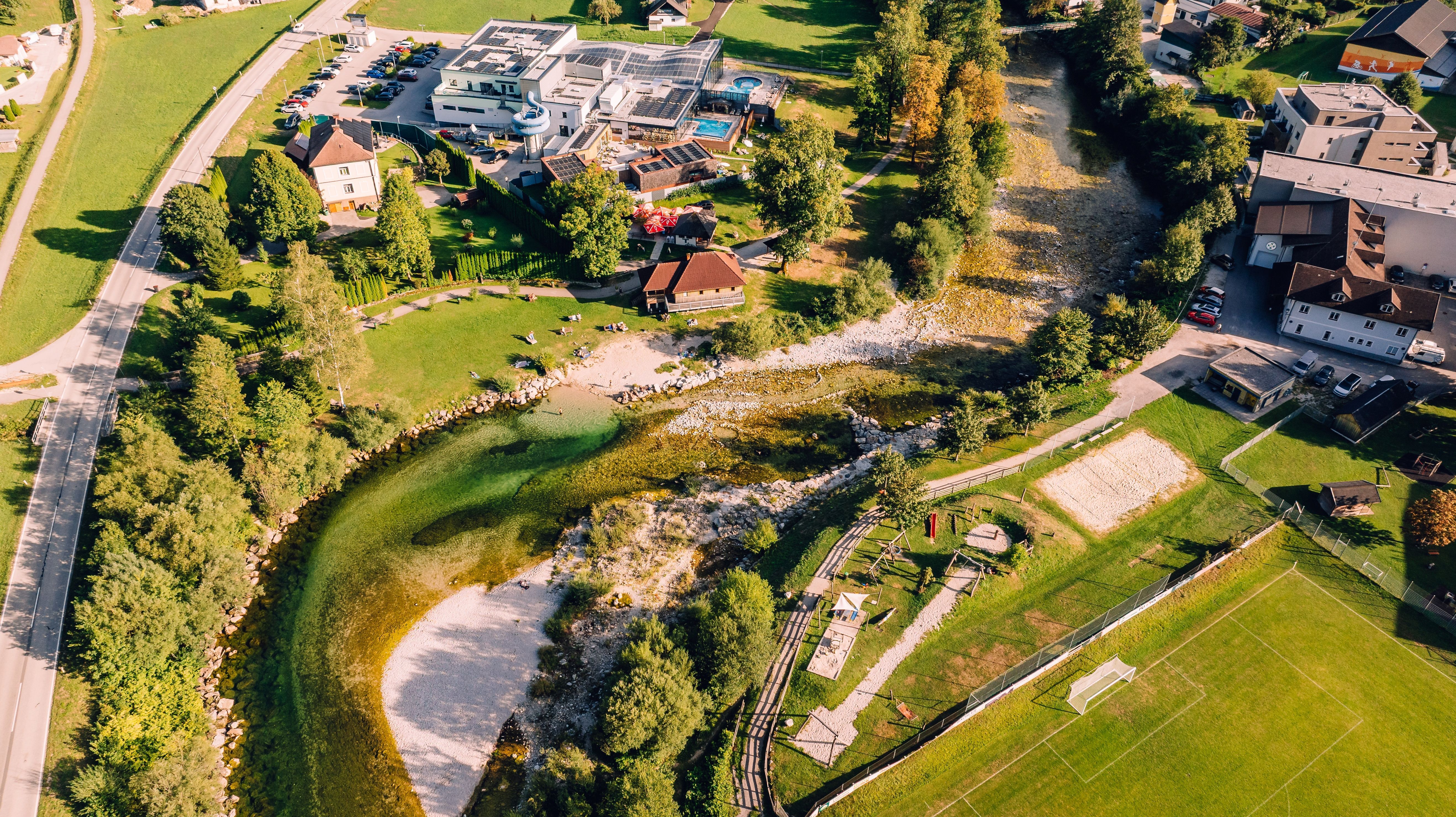 Aerial view of a riverbank with a hotel complex, parking lots, meadows, and a soccer field next to a shallow, rocky riverbed.