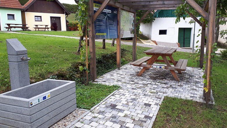 A rest area with a drinking fountain, wooden table and bench under a pergola, surrounded by green meadows and small buildings.