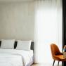 Modern hotel room with double bed, white sheets, wooden shelf and orange chair in front of a window with curtains.