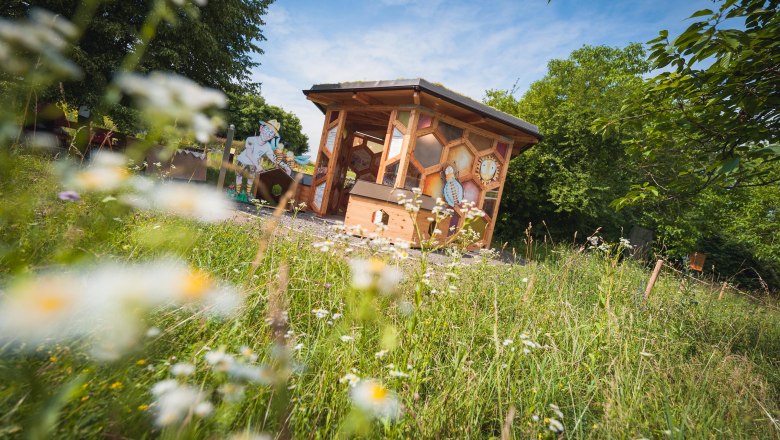 A wooden bee pavilion stands in a flowering meadow, surrounded by trees and a blue sky.
