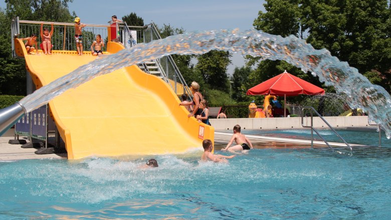 Children playing on a yellow water slide in an outdoor pool.