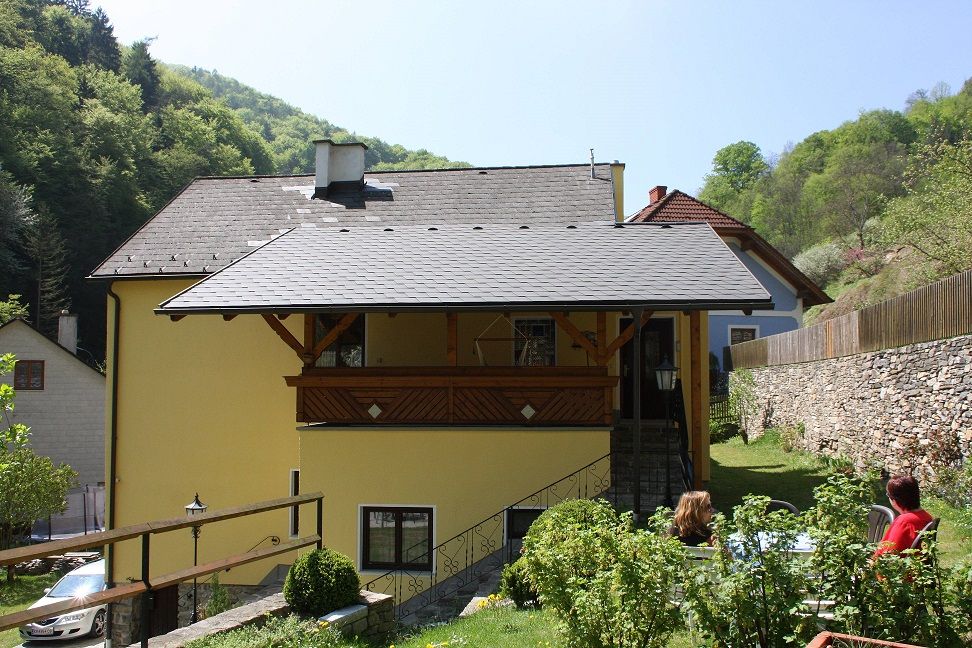 Yellow house with balcony, surrounded by green landscape and stone wall.