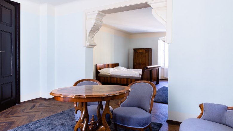 An elegant room with antique furniture, including a wooden table, two chairs and a bed in the background.