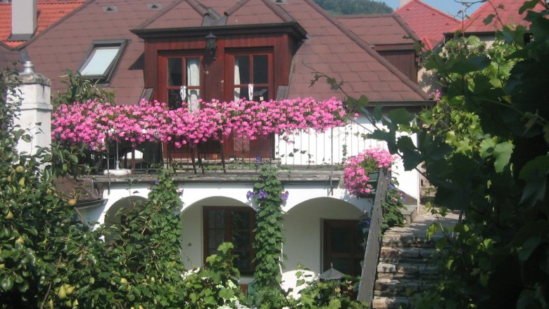 Guesthouse Haiminger, © Gästehaus Schmelz A guest house with a balcony full of pink flowers, surrounded by green vegetation.