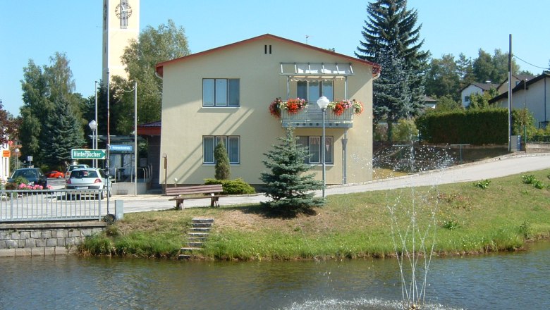 A yellow building with flower boxes and a bell tower in the background, with a pond and fountain in front of it.