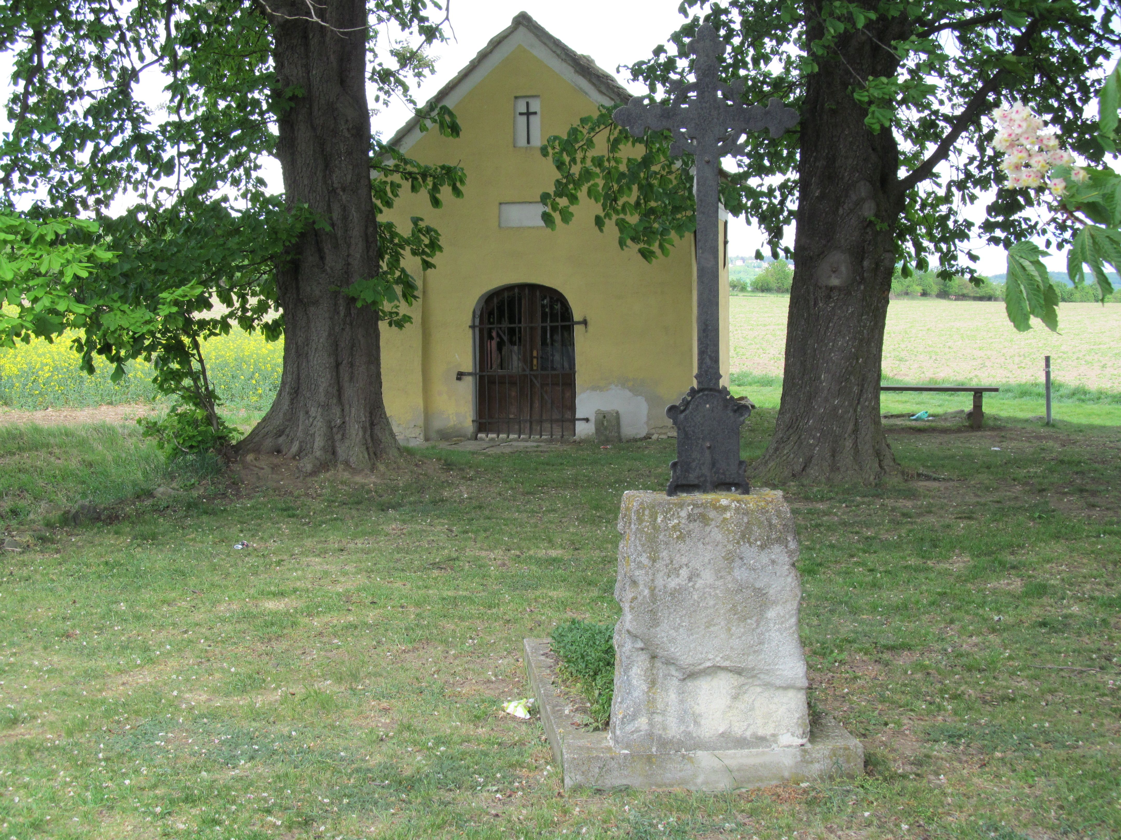 A yellow chapel with a cross in the foreground, surrounded by trees and meadows.