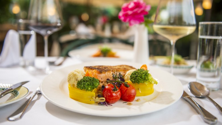 A plate of grilled salmon, broccoli, cauliflower, yellow peppers and cherry tomatoes, on a table set with wine glasses.