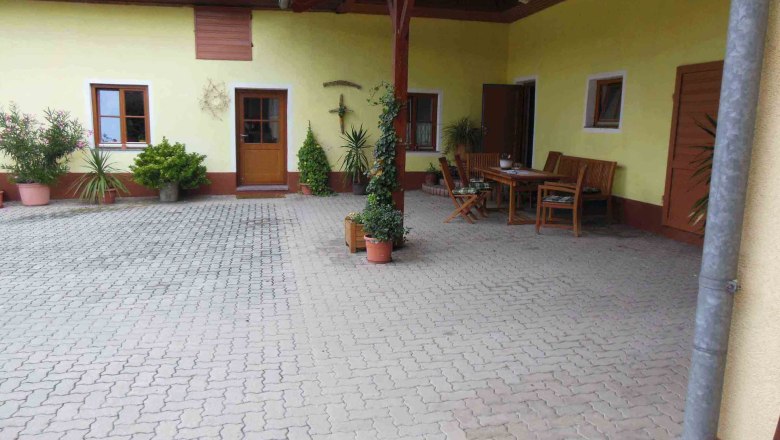 Vineyard view, © WEinbergblick Inner courtyard with yellow façade, wooden table and chairs, surrounded by plants in pots.