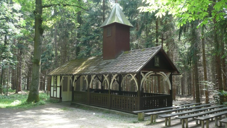 A small chapel in the forest with wooden benches outside.