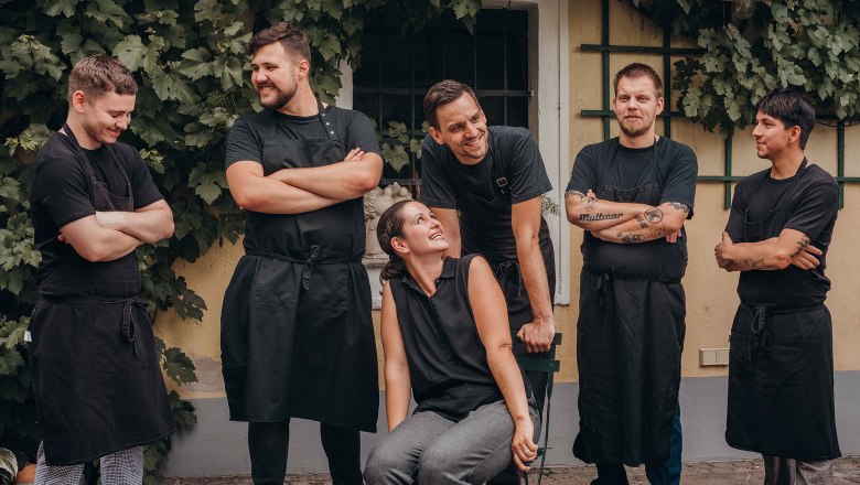 A group of six people in black work clothes stand and sit smiling in front of a wall overgrown with vines.