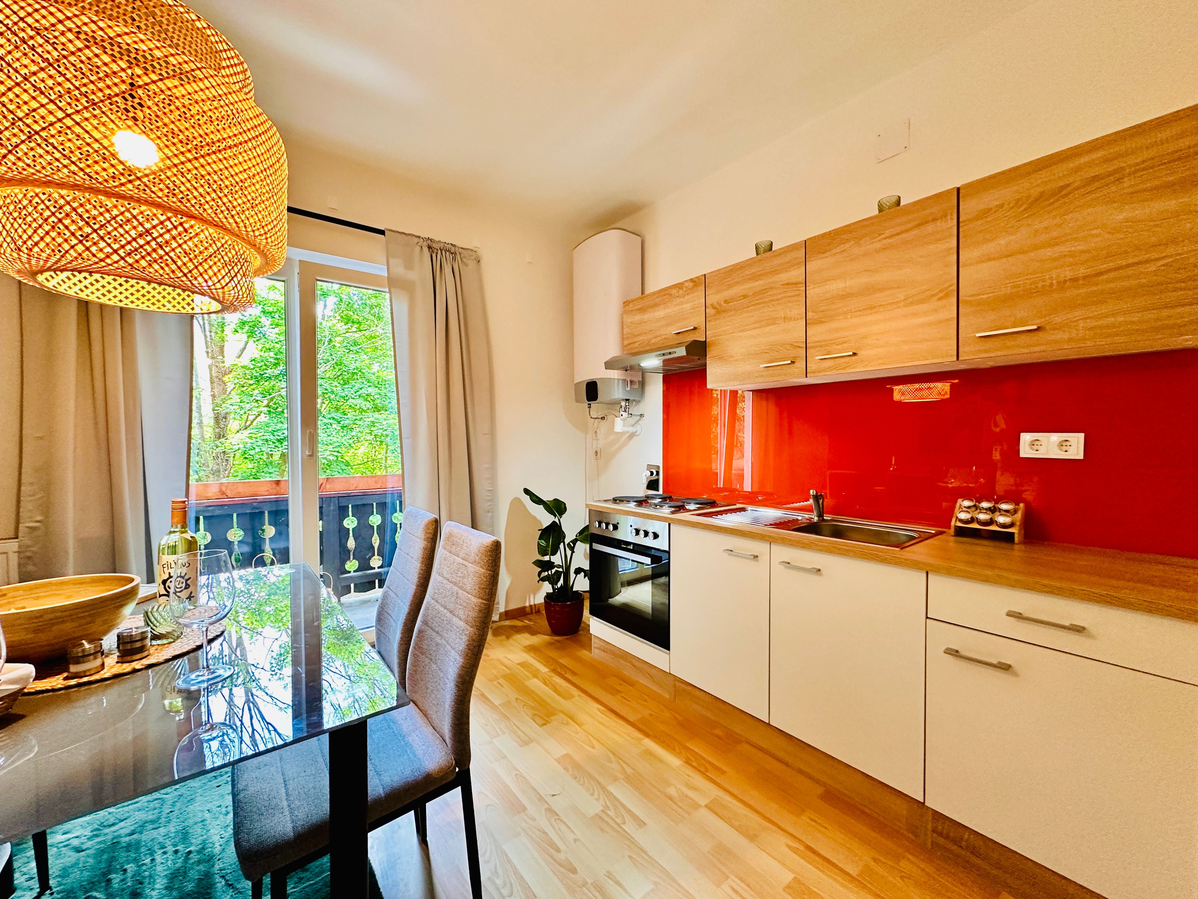 Modern kitchen with wooden units, red splashback and dining table in front of a window with a view of the greenery.