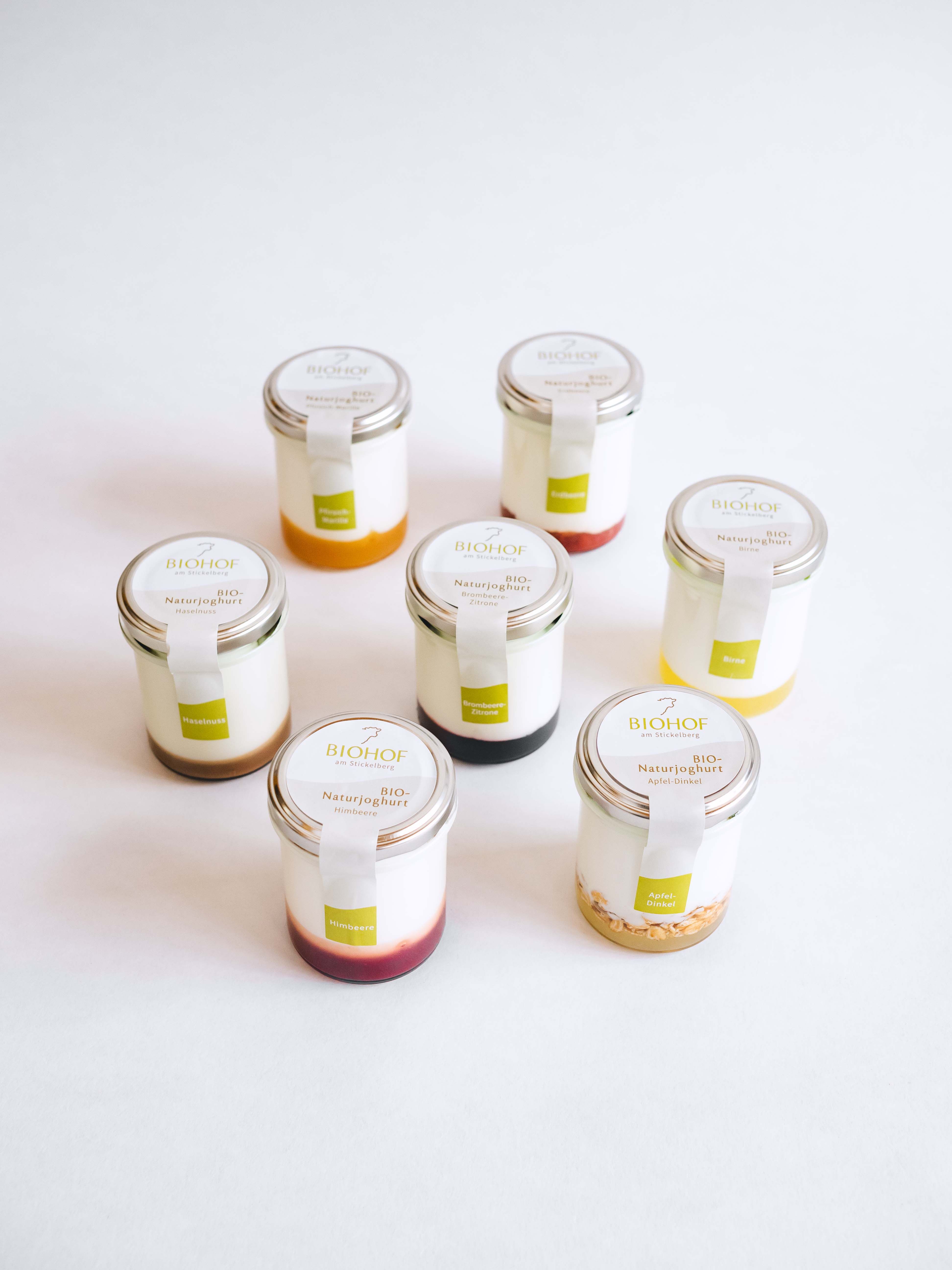 Seven glasses of natural yoghurt, each with different fruit toppings, against a white background.