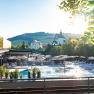 Outdoor area of a hotel with pool, parasols and sun loungers, surrounded by nature and a church in the background.