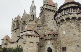 Two people are standing in front of Kreuzenstein Castle, surrounded by old stone walls and towers.
