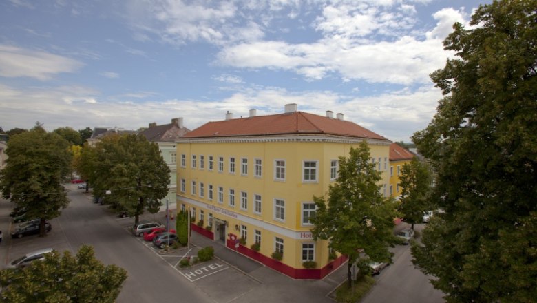 Exterior view of a yellow hotel with a red roof, surrounded by trees and streets.