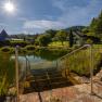 Steps lead to a pond with clear water, surrounded by green countryside and a house in the background, under a bright blue sky with sunshine.