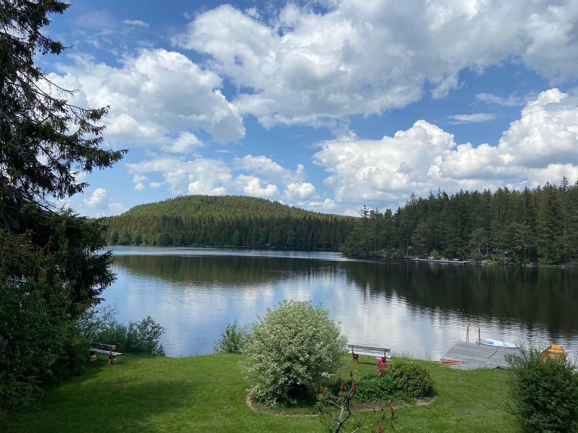 A calm lake with wooded shores and a jetty in the foreground, surrounded by a green landscape and under a cloudy sky.