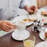 Person holding a coffee cup over a laid breakfast table with juice, bread rolls and jam.