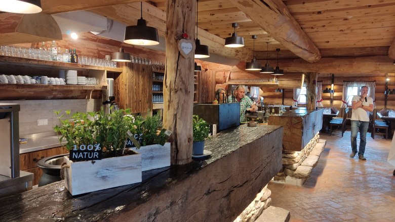 Interior view of a rustic mountain hut with wooden beams, bar and plants.