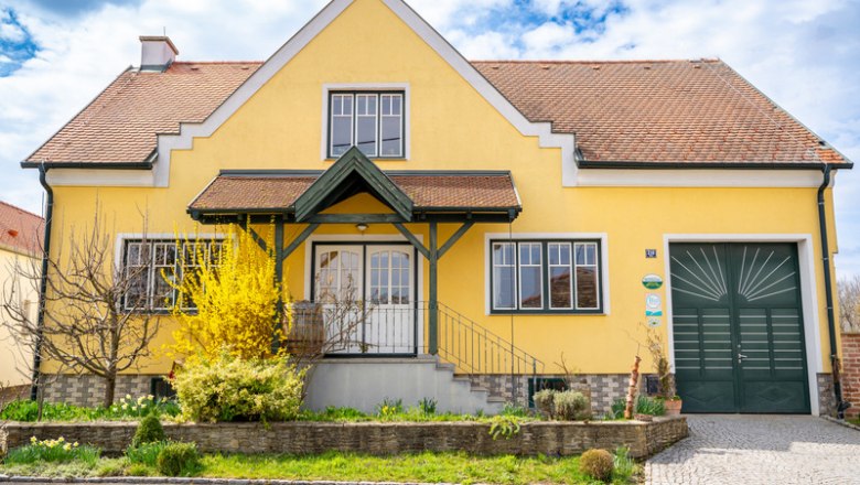 Yellow house with red roof and green gate, surrounded by garden and flowering shrubs.