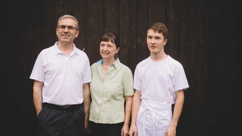 Three people stand in front of a dark wooden wall, smiling and looking into the camera.