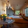 Interior view of a modern tree house lodge with wooden walls, bed, table with flowers and large windows with a view of the greenery.