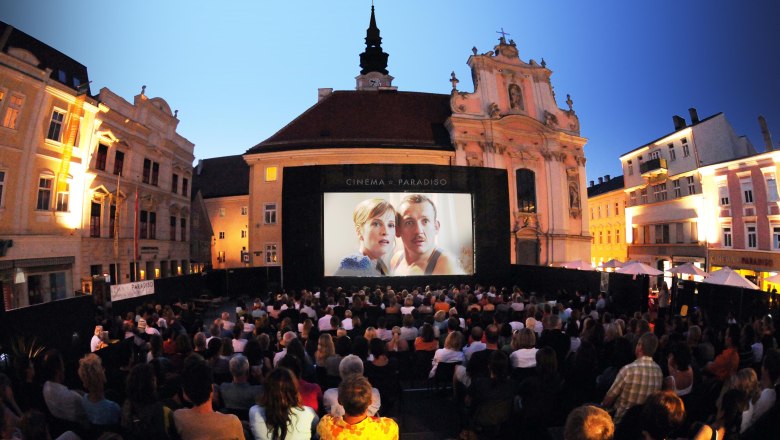 Open-air movie night in a square with a large audience in front of a big screen.