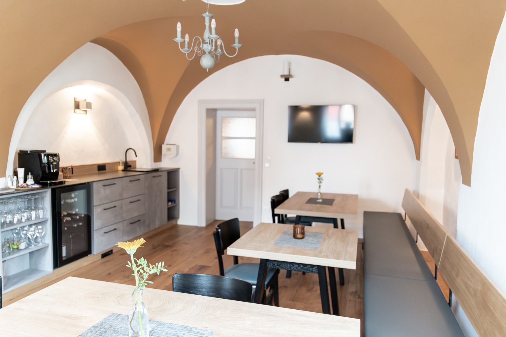 A cozy breakfast room with wooden tables, chairs and a bench. A TV hangs on the wall and there is a kitchenette with a coffee machine.