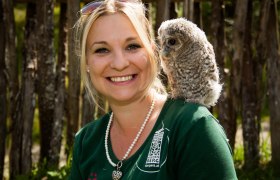 A woman laughs into the camera while an owl sits quietly on her shoulder.