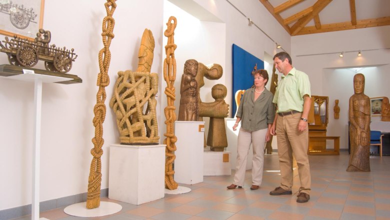 Two people look at wooden sculptures in an exhibition room with a wooden beamed ceiling.