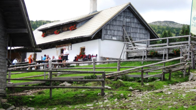 Alpine hut with people in a meadow, surrounded by wooden fences.