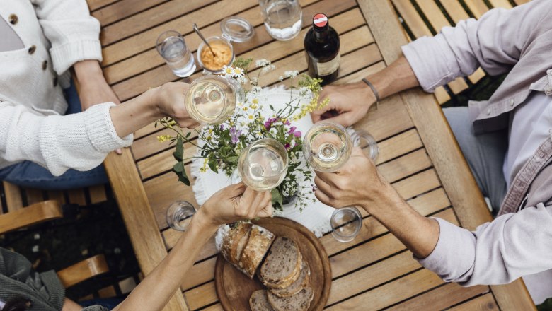 Three people clink glasses of wine at a wooden table with bread and flowers on it.