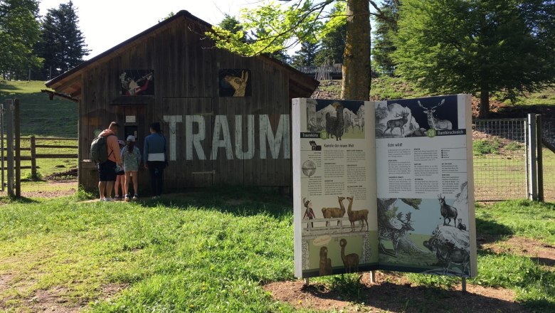 A family stands in front of a wooden hut with the inscription 'TRAUM'. Next to it is an information board about animals in a green park.