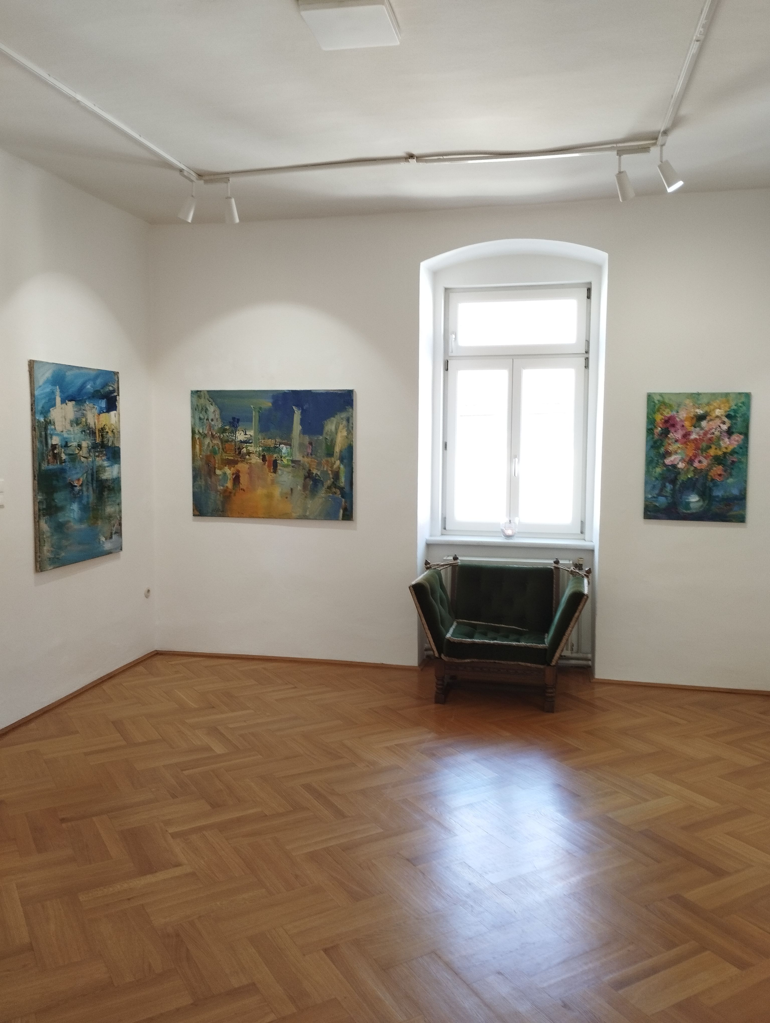 Interior view of a gallery with three paintings on the walls and a green armchair in front of a window.