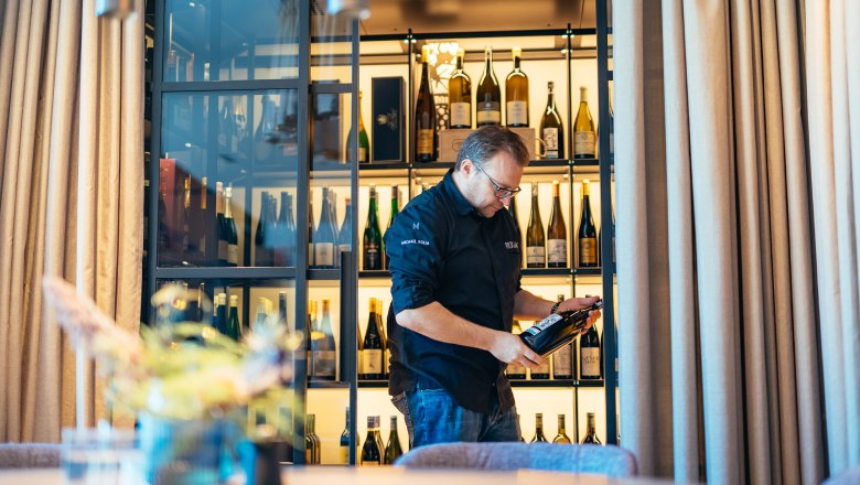A man looks at a bottle of wine in a walk-in wine cabinet with many bottles.