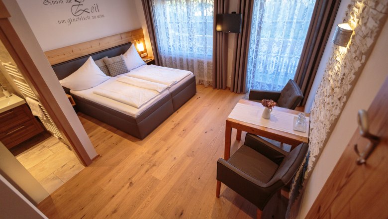 Cozy double room with wooden floor, double bed, small table and chairs, decorative curtains and a TV on the wall.