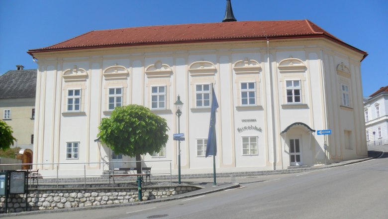Historic building with a red roof and white walls, surrounded by trees and a road.