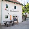 Exterior view of a building with the inscription 'Hauser Chalet Weingenuss'. In front of the building is a wooden table and a wine barrel.