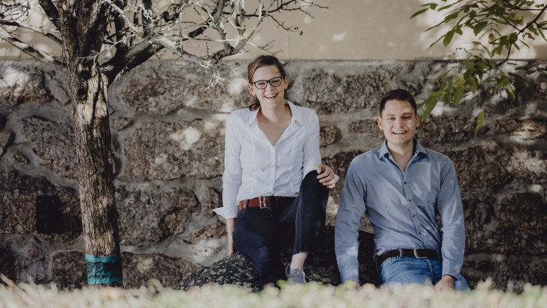 Two people sit smiling on a stone in front of a stone wall, surrounded by trees.