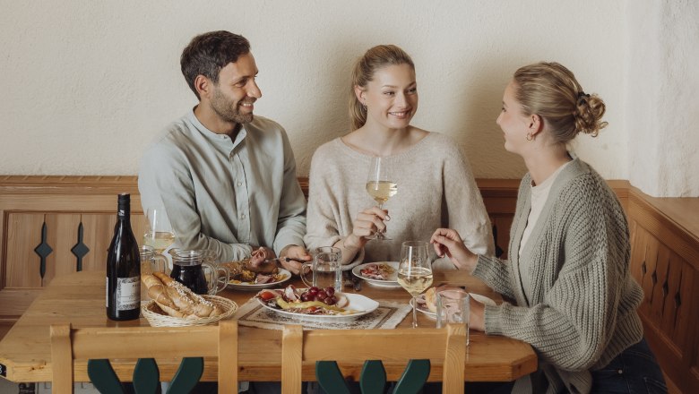Three people sit at a wooden table in a wine cellar and enjoy wine and food.