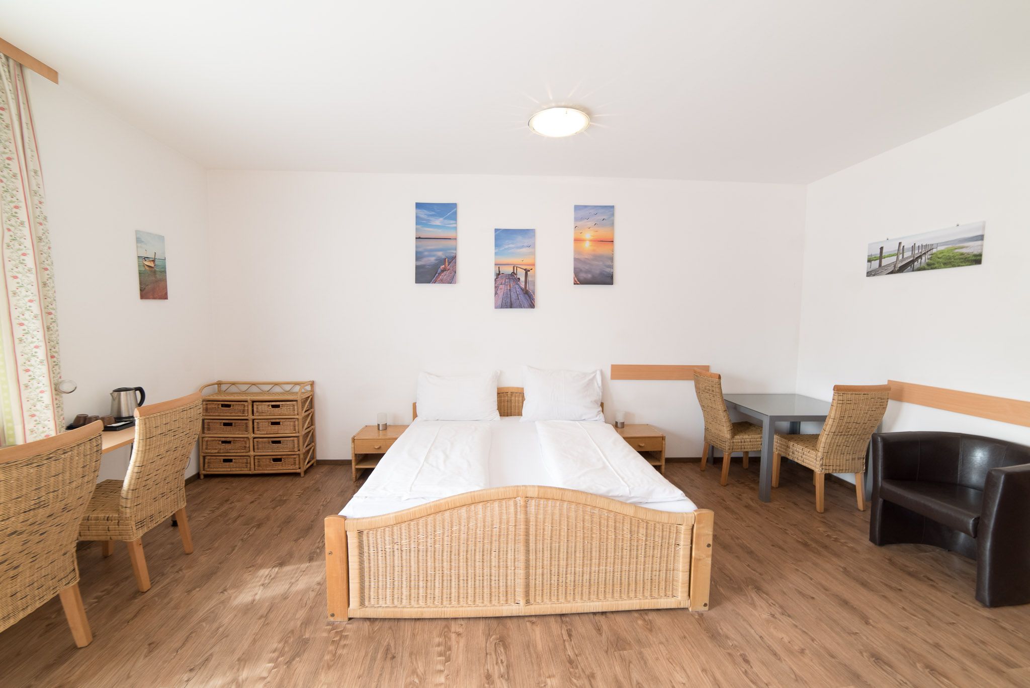 Large bedroom with light beige tones and rattan furniture. Sitting area, desk, double bed, couch, bedside cabinet.