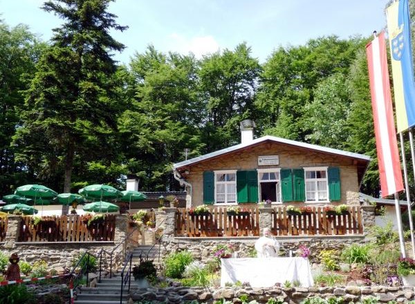 A mountain hut with green shutters, surrounded by trees, with a garden and parasols in the foreground.