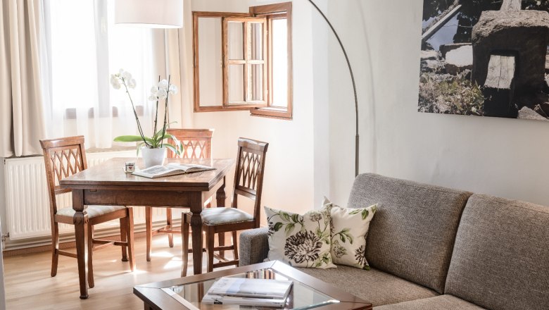 Cozy living room with grey sofa, wooden table and chairs, bright window and plants.