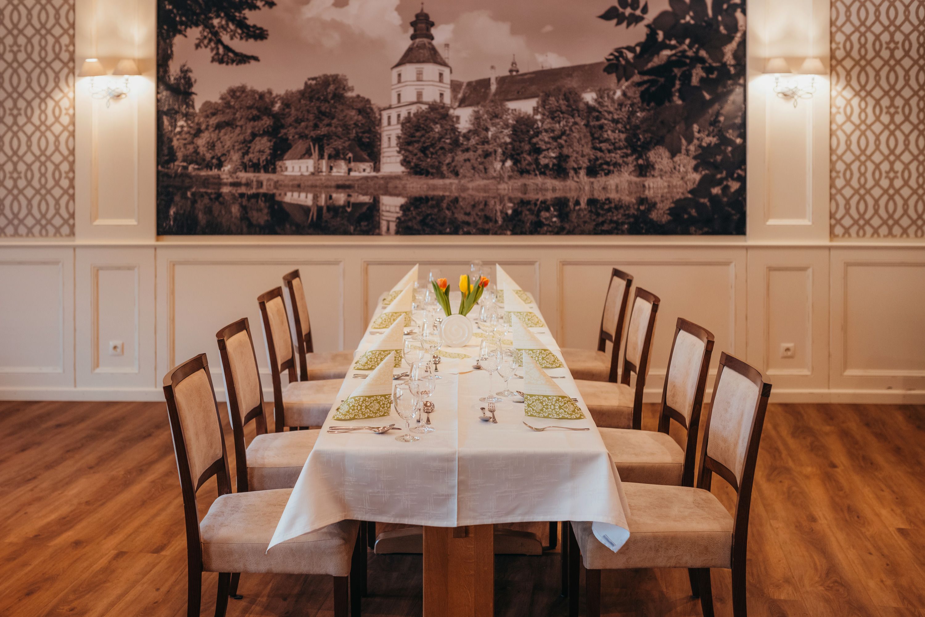 Elegant dining room with a long table, laid with white tablecloths and green napkins, with a large picture of the castle in the background.