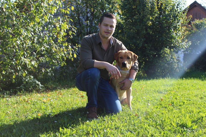 A man kneels in a meadow next to a dog, surrounded by trees and sunlight.
