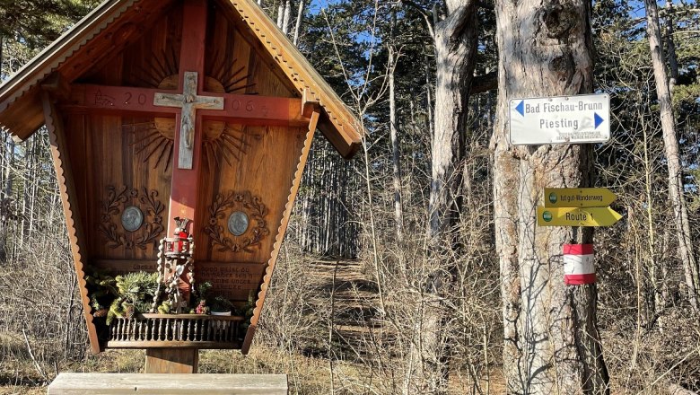 Wooden cross with bench in the forest, next to it a signpost with directions.