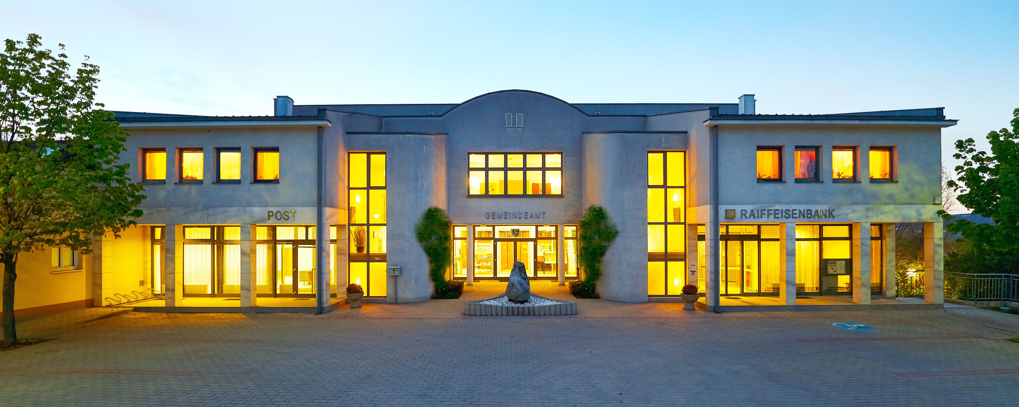 Illuminated building with the inscription 'Gemeindeamt', 'Post' and 'Raiffeisenbank'.