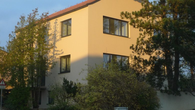 A modern, three-storey house with a yellow fa&ccedil;ade and red roof tiles, surrounded by trees and shrubs.