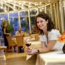 A woman in a conservatory restaurant holds a glass of red wine. Other guests and a waitress can be seen in the background.